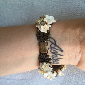 May include: A gold-tone bracelet with a floral design. The bracelet is adorned with white flowers and pearls. The bracelet has a tag that reads "Colleen Ireland".