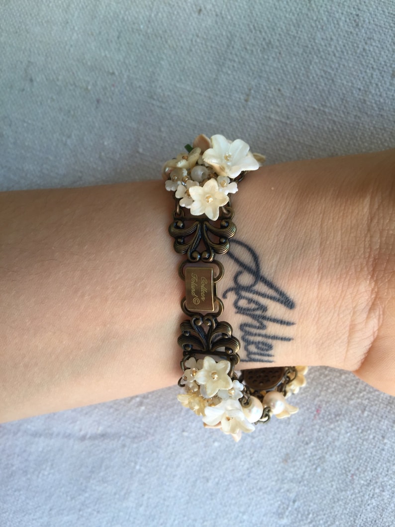 May include: A gold-tone bracelet with a floral design. The bracelet is adorned with white flowers and pearls. The bracelet has a tag that reads "Colleen Ireland".