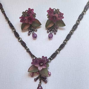 Pu&ograve; includere: Un set di collana e orecchini abbinati con un design floreale. I gioielli hanno una catena in bronzo scuro e sono adornati con grappoli di fiori viola, foglie verdi e perline viola pendenti. Gli orecchini sono a clip.