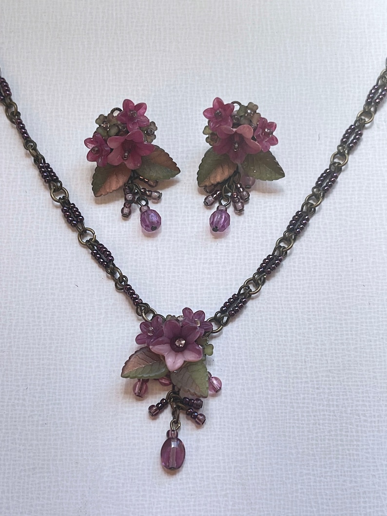 Pu&ograve; includere: Un set di collana e orecchini abbinati con un design floreale. I gioielli hanno una catena in bronzo scuro e sono adornati con grappoli di fiori viola, foglie verdi e perline viola pendenti. Gli orecchini sono a clip.