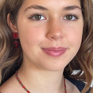 May include: A woman wearing a red beaded necklace with a dangling red flower charm. She is also wearing red beaded earrings.