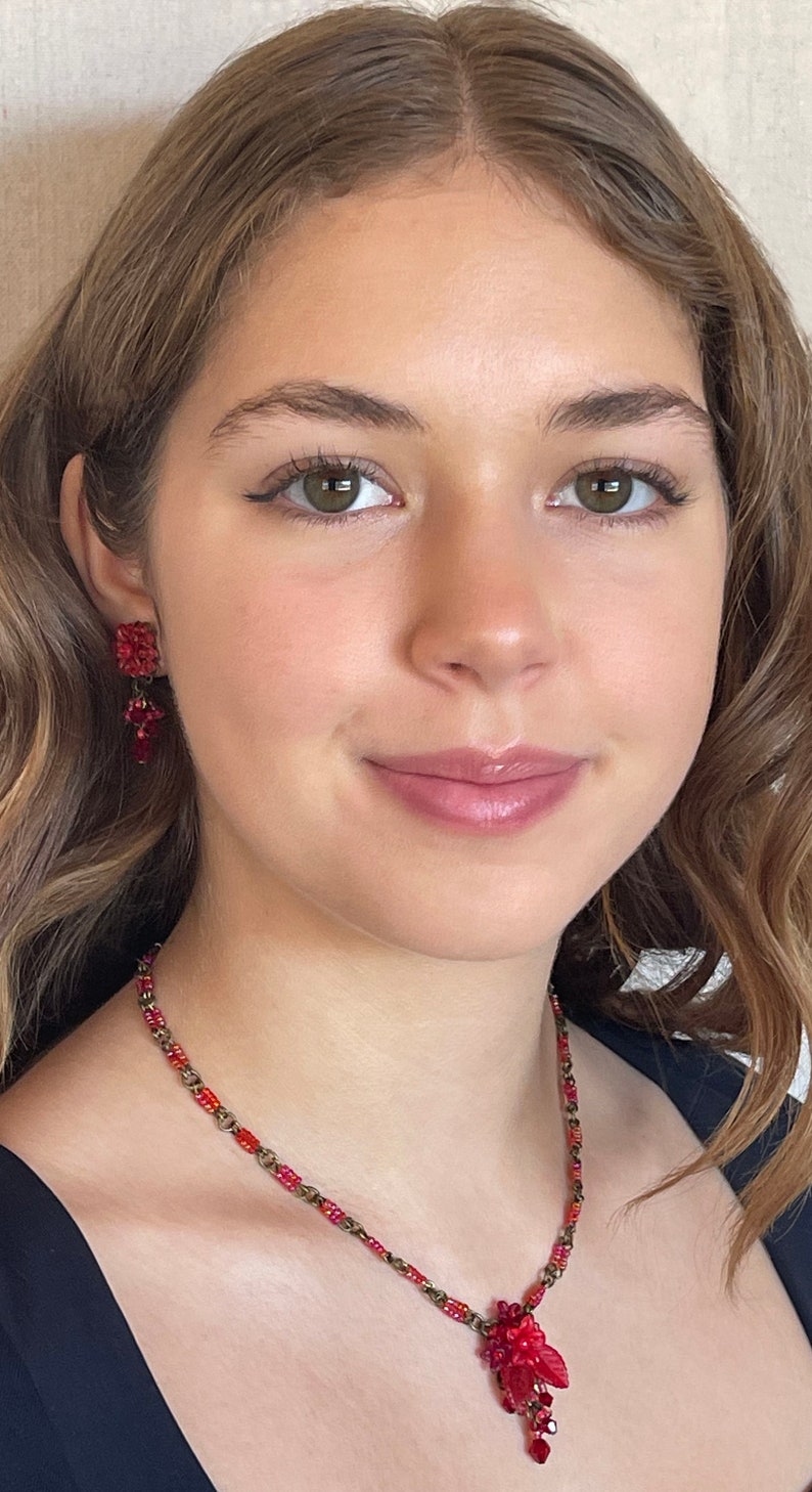 May include: A woman wearing a red beaded necklace with a dangling red flower charm. She is also wearing red beaded earrings.