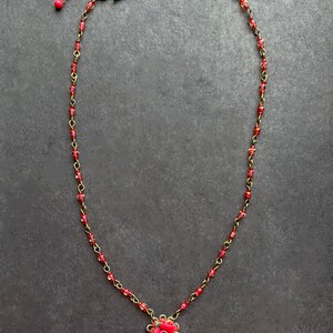 Może przedstawiać: Naszyjnik z łańcuszkiem w kolorze brązu, przeplatanym małymi czerwonymi koralikami. Zawieszka składa się z rzeźbionej czerwono-r&oacute;żowej r&oacute;ży na ażurowej podstawie z brązu, z pojedynczym wiszącym czerwonym koralikiem u dołu i zapięciem na haczyk.
