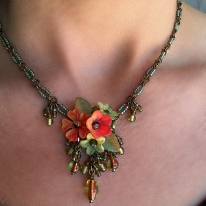 Könnte beinhalten: Eine Halskette mit einer Bronzekette und einem Anhänger mit orangefarbenen und roten Blumen. Die Halskette hat grüne Blätter und hängende Perlen in Grün- und Bernsteintönen. Die Halskette wird um den Hals getragen.