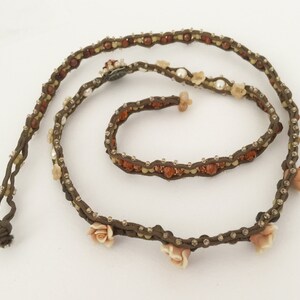 May include: A brown beaded necklace with small, clear crystals and amber-colored beads. The necklace features small, light pink rose accents and a knotted end. The jewelry is displayed on a white background.