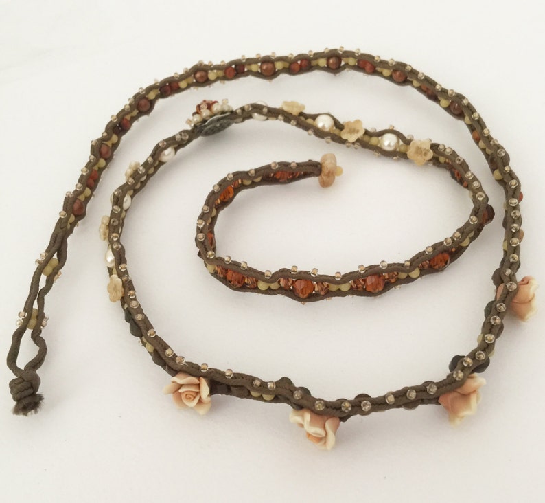 May include: A brown beaded necklace with small, clear crystals and amber-colored beads. The necklace features small, light pink rose accents and a knotted end. The jewelry is displayed on a white background.