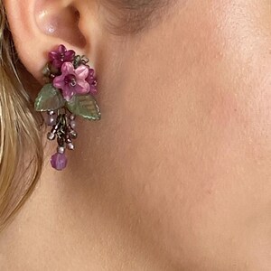 Pu&ograve; includere: Un orecchino floreale con grappoli di fiori rosa e viola, foglie verdi e perline pendenti. L'orecchino &egrave; attaccato all'orecchio. Il design &egrave; intricato e dettagliato, con un'estetica vintage.