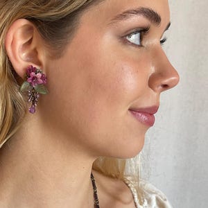 Pu&ograve; includere: Primo piano di un orecchino floreale. L'orecchino presenta grappoli di fiori rosa e viola, foglie verdi e perline viola pendenti. L'orecchino &egrave; indossato sull'orecchio di una persona.