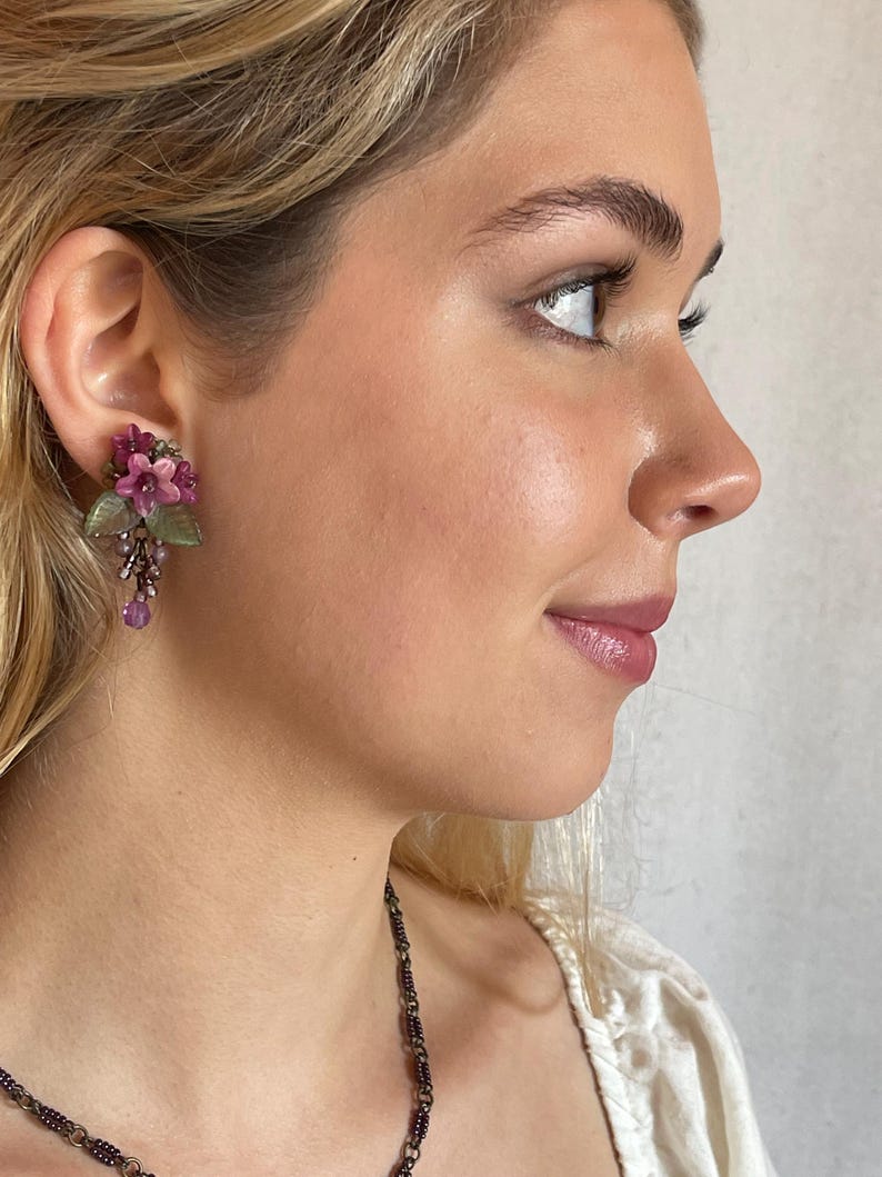 Pu&ograve; includere: Primo piano di un orecchino floreale. L'orecchino presenta grappoli di fiori rosa e viola, foglie verdi e perline viola pendenti. L'orecchino &egrave; indossato sull'orecchio di una persona.
