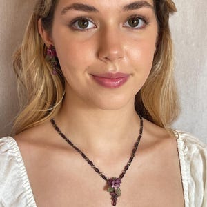 Pu&ograve; includere: Un set di gioielli floreali con collana e orecchini. La collana ha una catena di perline scure con un gruppo di fiori viola e una perla a forma di goccia. Gli orecchini rispecchiano il design floreale.