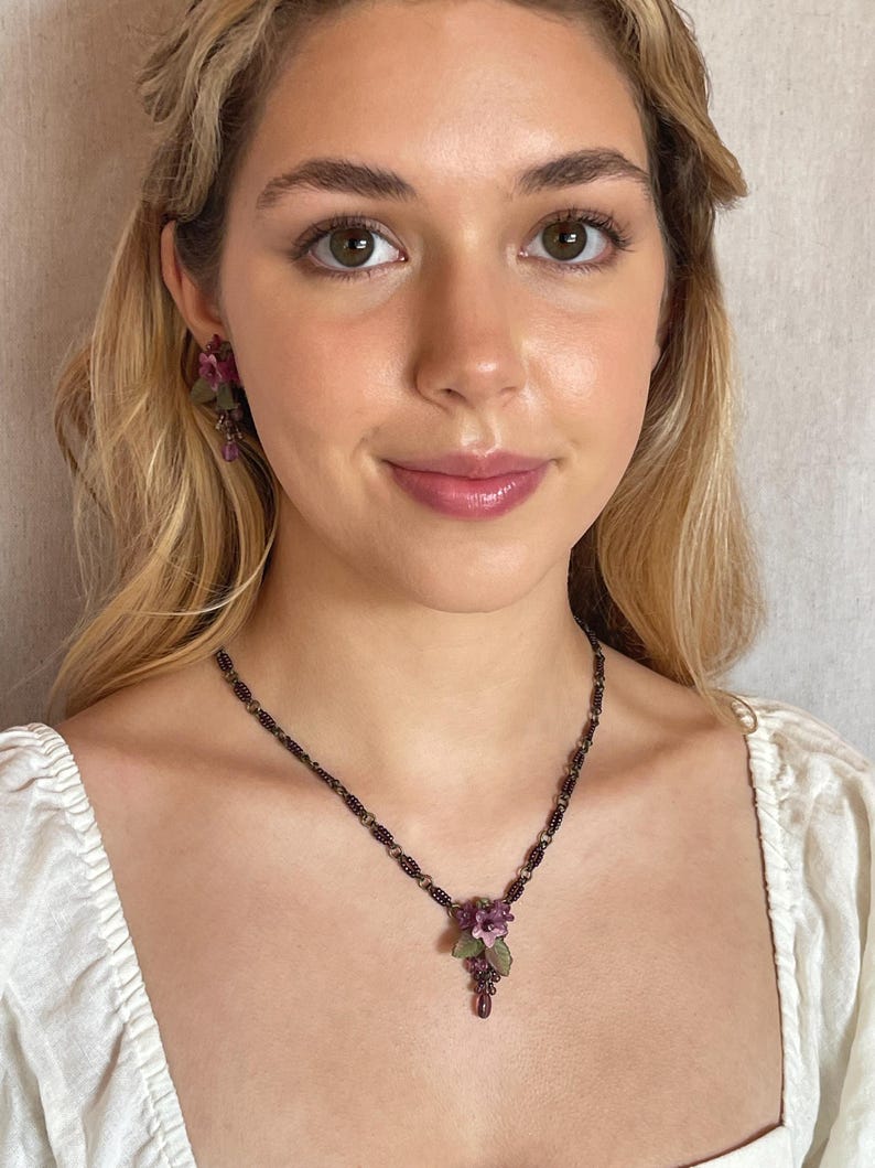 Pu&ograve; includere: Un set di gioielli floreali con collana e orecchini. La collana ha una catena di perline scure con un gruppo di fiori viola e una perla a forma di goccia. Gli orecchini rispecchiano il design floreale.