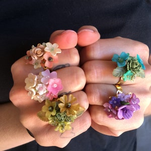 以下が含まれることがあります： 花柄のデザインが施された金の指輪が5つ。それぞれの指輪には、ピンク、ブルー、グリーン、パープルなど、カラフルな花が集まっています。