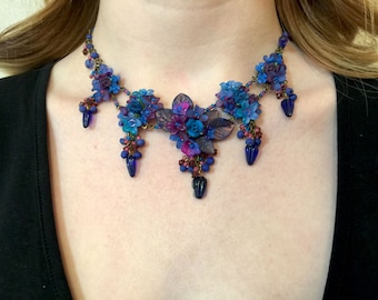 Può includere: Una collana delicata con un designo floreale con fiori blu, viola e rosa. La collana è realizzata con perline di metallo e vetro e ha un effetto a cascata.