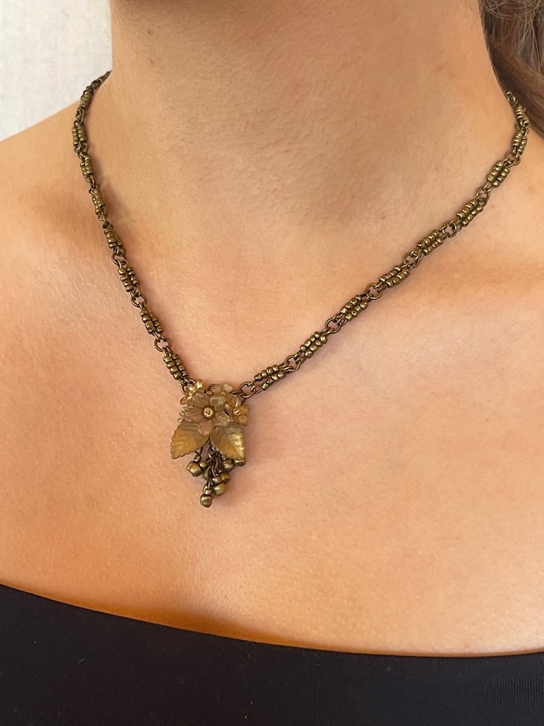 May include: Vintage brass-coloured necklace with a floral pendant, leaves, and grape-like clusters. The chain is made of small, connected links. The necklace is worn around the neck.