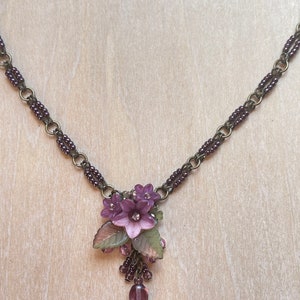 Pu&ograve; includere: Una delicata collana con un grappolo di fiori viola con un strass centrale, accentuato da foglie verdi e rosa. La collana ha una catena in bronzo scuro con accenti di perline viola e una perla a forma di goccia.