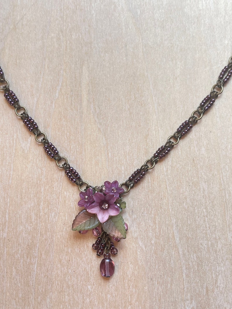 Pu&ograve; includere: Una delicata collana con un grappolo di fiori viola con un strass centrale, accentuato da foglie verdi e rosa. La collana ha una catena in bronzo scuro con accenti di perline viola e una perla a forma di goccia.