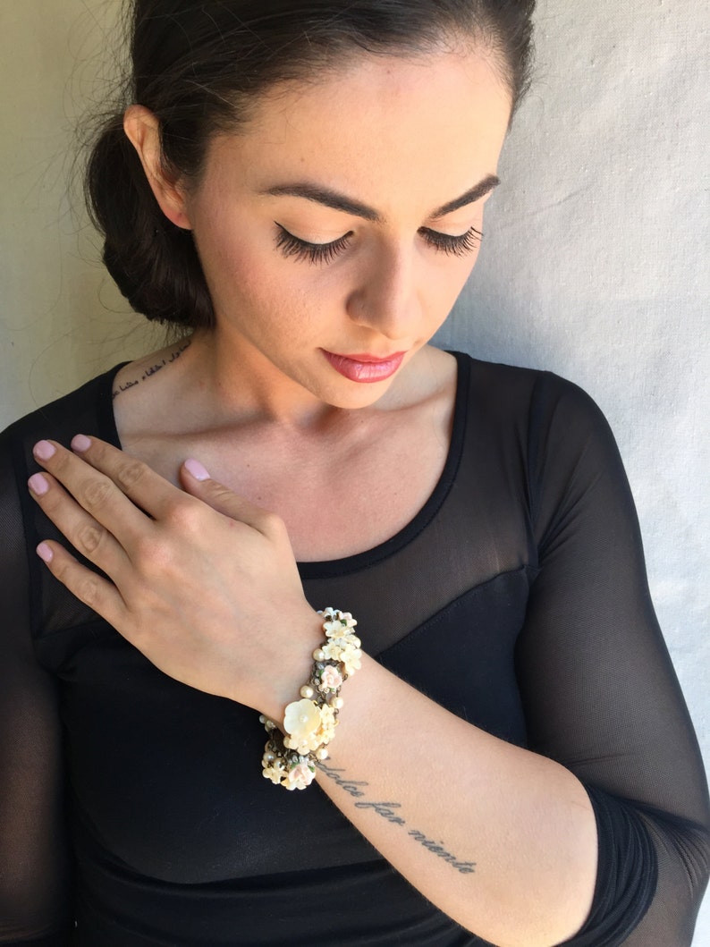 May include: A close-up of a woman's arm wearing a white and pink floral bracelet. The bracelet is made of small flowers and pearls. The woman has a tattoo on her arm that says "sola far niente".