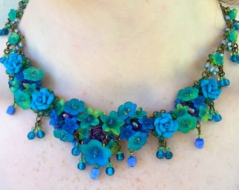 Può includere: Una collana floreale blu e verde con una catena di bronzo e perline blu e verdi. La collana è realizzata in pasta polimerica e presenta una varietà di forme di fiori.