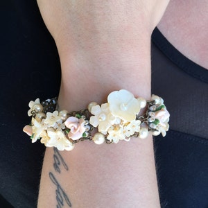 May include: A white and pink floral bracelet with pearl accents. The bracelet is made of metal and has a delicate, feminine design.