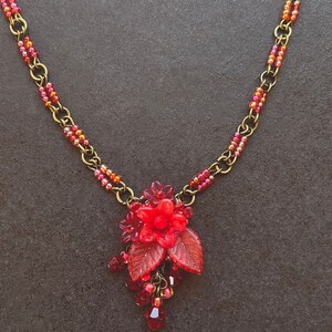 May include: A red beaded necklace with a red flower pendant and leaves. The necklace is made with small red and gold beads and has a brass chain.