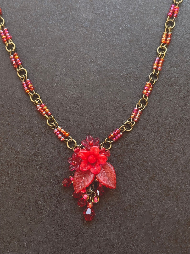 May include: A red beaded necklace with a red flower pendant and leaves. The necklace is made with small red and gold beads and has a brass chain.