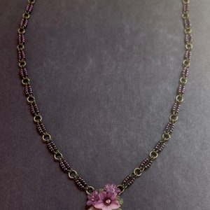 Pu&ograve; includere: Una delicata collana con un ciondolo floreale. La collana ha una catena color bronzo con perline viola e un ciondolo con fiori viola, foglie verdi e un cristallo centrale. La collana &egrave; lunga circa 46 cm.