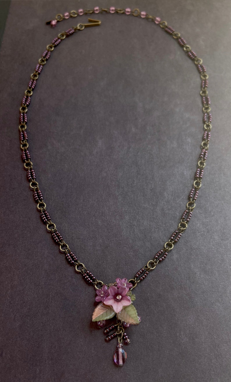 Pu&ograve; includere: Una delicata collana con un ciondolo floreale. La collana ha una catena color bronzo con perline viola e un ciondolo con fiori viola, foglie verdi e un cristallo centrale. La collana &egrave; lunga circa 46 cm.