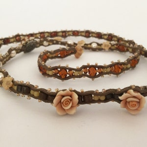 May include: A brown beaded necklace and bracelet set. The necklace features small, light-colored beads, amber-colored crystals, and three peach-colored rose accents. The bracelet has a similar design. The set is made of a brown cord.