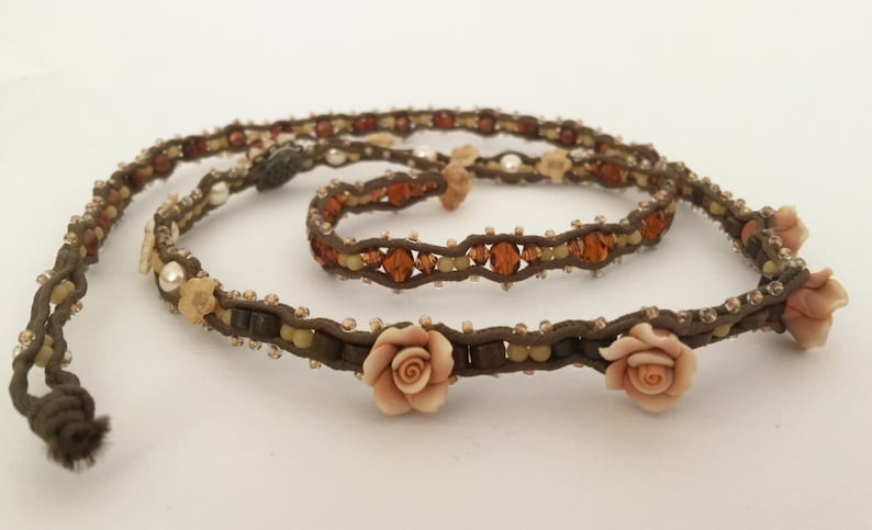 May include: A brown beaded necklace and bracelet set. The necklace features small, light-colored beads, amber-colored crystals, and three peach-colored rose accents. The bracelet has a similar design. The set is made of a brown cord.