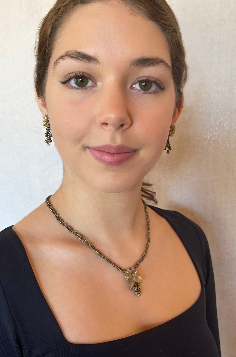 May include: A vintage jewellery set featuring a necklace and matching earrings. The necklace has a delicate chain with a floral pendant. The earrings are dangling, with a similar floral design. The jewellery is a muted gold colour.