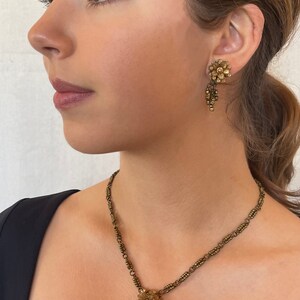 May include: Vintage gold-toned jewellery set comprising a necklace and earrings. The necklace features a floral pendant with a cluster of grapes. The earrings have a similar floral design with dangling beads. The jewellery is worn by a person.