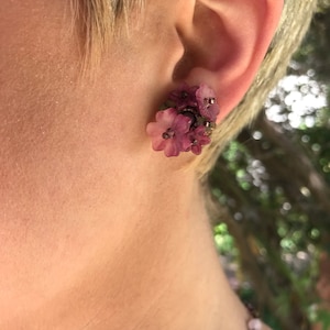 Puede incluir: Un par de pendientes de color rosa en forma de flor con un racimo de pequeñas cuentas moradas. Los pendientes se llevan en la oreja de una persona con el pelo corto y rubio.