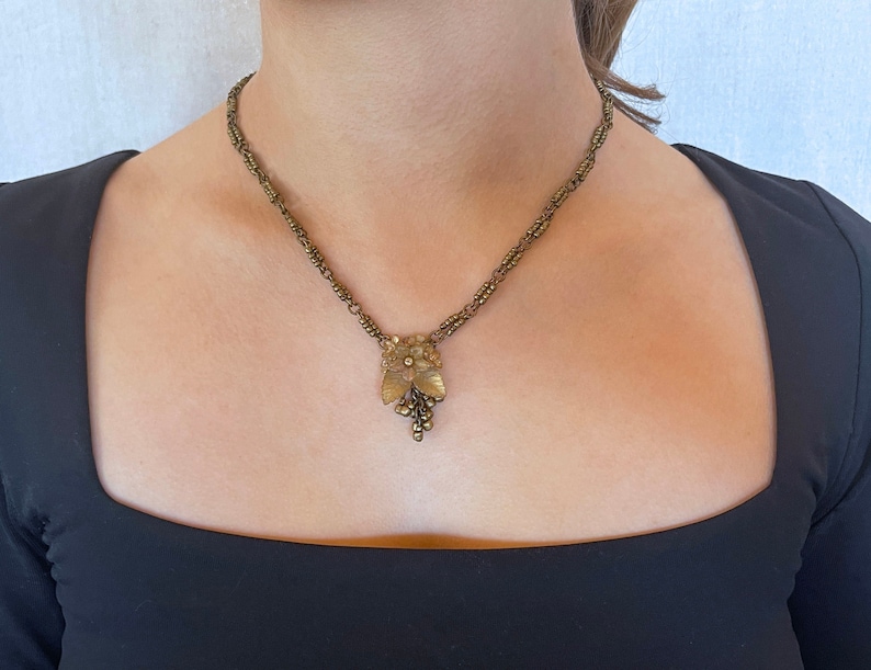 May include: Vintage brass-coloured necklace with a floral pendant and grape cluster. The necklace has a delicate chain design. The pendant has intricate detailing and is worn against a black top.