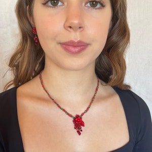May include: A red beaded necklace with a floral pendant. The pendant is made of red beads and has a gold-tone finish. The necklace is worn by a person with long brown hair.