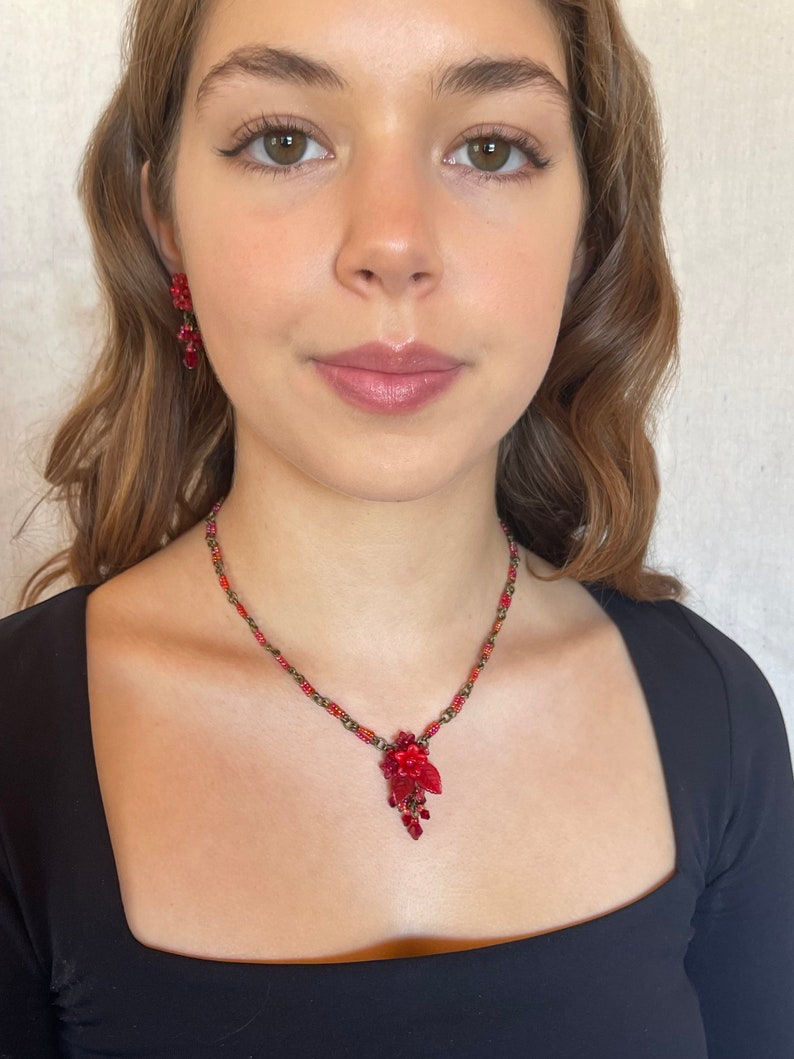 May include: A red beaded necklace with a floral pendant. The pendant is made of red beads and has a gold-tone finish. The necklace is worn by a person with long brown hair.