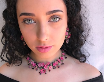 May include: A woman wearing a black off-the-shoulder top with a pink floral choker necklace and matching earrings.