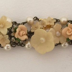 May include: A delicate bracelet featuring a chain with a floral design. The bracelet is adorned with small white pearls and pastel pink flowers. The clasp is made of metal.
