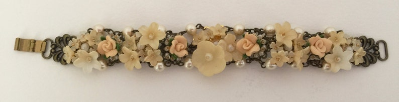 May include: A delicate bracelet featuring a chain with a floral design. The bracelet is adorned with small white pearls and pastel pink flowers. The clasp is made of metal.