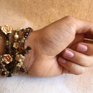 May include: A multi-strand wrap bracelet featuring brown cord, cream-colored pearls, and small beads. The bracelet is adorned with small, light-colored fabric flowers and is wrapped around a wrist.