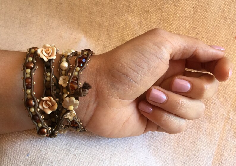 May include: A multi-strand wrap bracelet featuring brown cord, cream-colored pearls, and small beads. The bracelet is adorned with small, light-colored fabric flowers and is wrapped around a wrist.