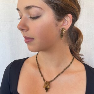 May include: A vintage jewellery set comprising a necklace and earrings. The necklace features a delicate chain with a floral pendant and a cluster of small beads. The earrings have a similar floral design. The set is in a bronze tone.