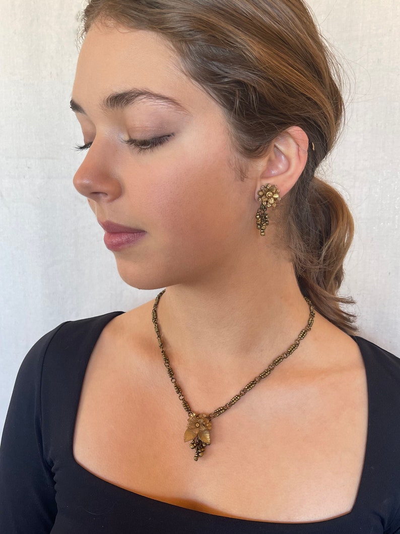 May include: A vintage jewellery set comprising a necklace and earrings. The necklace features a delicate chain with a floral pendant and a cluster of small beads. The earrings have a similar floral design. The set is in a bronze tone.