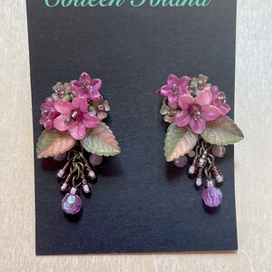 Pu&ograve; includere: Un paio di orecchini floreali con fiori rosa, foglie verdi e color rame e perline viola pendenti. Gli orecchini sono esposti su una carta nera con il testo "Colleen Toland" in bianco.