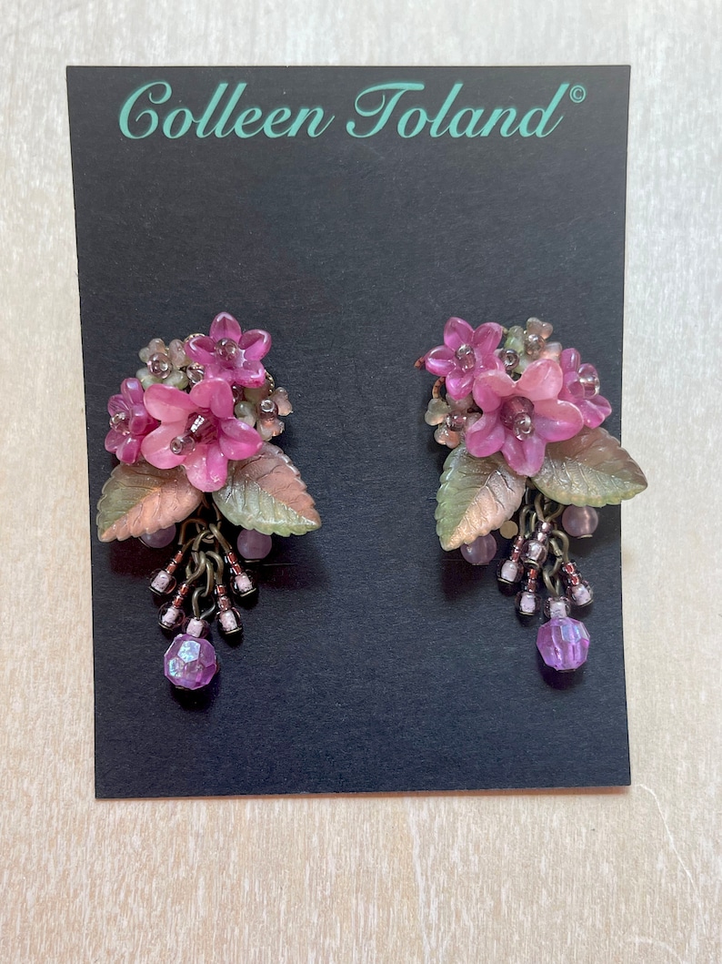 Pu&ograve; includere: Un paio di orecchini floreali con fiori rosa, foglie verdi e color rame e perline viola pendenti. Gli orecchini sono esposti su una carta nera con il testo "Colleen Toland" in bianco.
