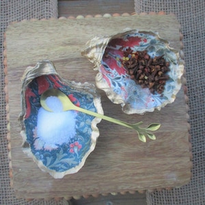 May include: Two decorative seashell-shaped bowls, one filled with white salt and a gold spoon, the other filled with brown cloves and red peppercorns. The bowls are on a wooden surface.
