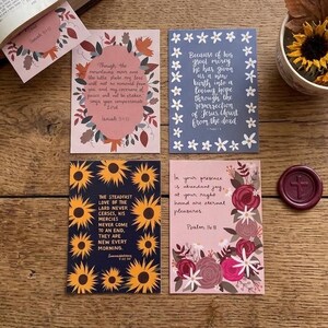 May include: Four rectangular cards featuring floral borders and printed scripture verses, a large open book with a bookmark, a ceramic mug filled with sunflowers and dried leaves, a circular wax seal, and two pens with pink and black accents.