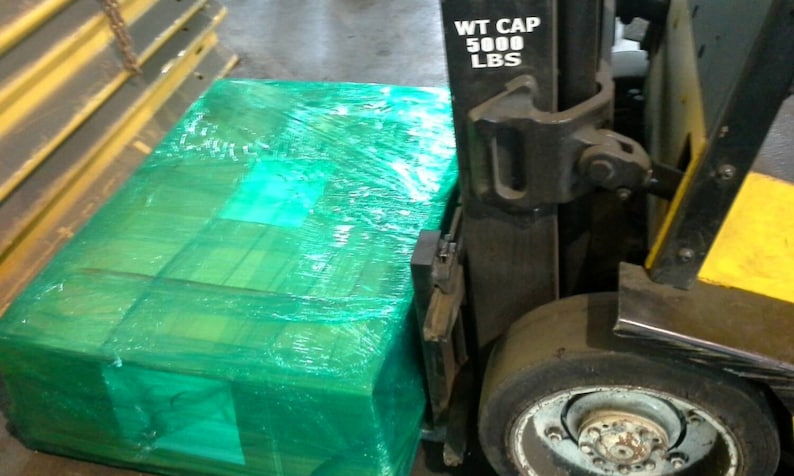 May include: A green pallet wrapped in plastic shrink wrap is being loaded onto a forklift. The forklift has a weight capacity of 5000 lbs.