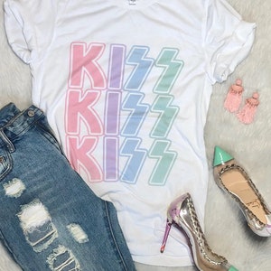 Puede incluir: Una camiseta blanca con la palabra "KISS" repetida tres veces en un degradado arco&iacute;ris. La camiseta se combina con unos vaqueros desgastados y zapatos de tac&oacute;n alto.