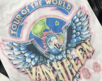 Puede incluir: Camiseta blanca con un gráfico vintage de Van Halen "Tour of the World 1984" que presenta un águila azul y blanca con un contorno rosa. El águila sostiene un globo terráqueo en sus garras.