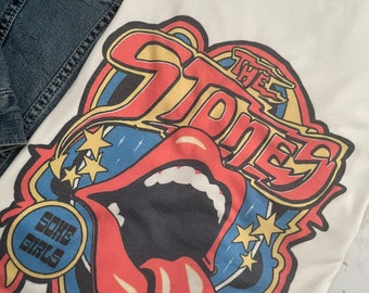 May include: White t-shirt with a graphic print of "The Stones" in a retro style. The graphic features a red tongue with a mouth, stars, and the text "Some Girls".